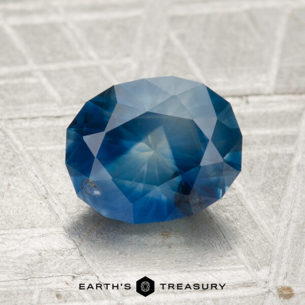 2.19-Carat Montana Sapphire (Heated) A 2.19-carat heated Montana sapphire, faceted in an oval shape, is displayed on a textured gray surface with the text "EARTH'S TREASURY" at the bottom.