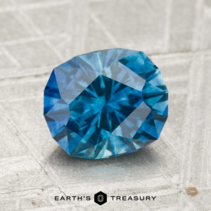 A faceted 2.06-carat heated Montana sapphire is displayed on a light textured surface, with the text "EARTH'S TREASURY" at the bottom.