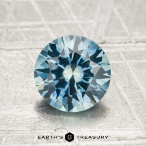 A 1.67-carat heated Montana sapphire, cut in a round brilliant shape, is displayed on a textured gray surface with the "Earth's Treasury" logo at the bottom.