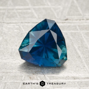 A 1.46-carat, heated Montana sapphire in trillion cut rests on a textured silver-gray surface, with "Earth's Treasury" displayed below.