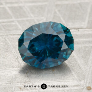A 2.17-carat heated Montana sapphire rests on a light textured surface, with "EARTH'S TREASURY" and a hexagon logo displayed beneath it.