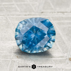 A 2.01-carat oval-cut Montana sapphire (heated) rests on a light textured surface, with "Earth's Treasury" printed below.