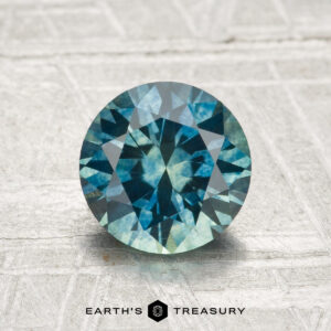 A round brilliant-cut 1.64-carat heated Montana sapphire in blue-green, displayed on a textured light surface, with "EARTH'S TREASURY" text at the bottom.