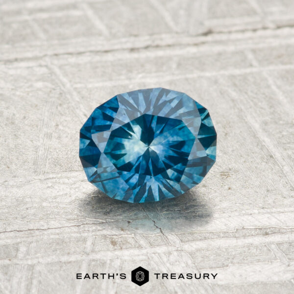 0.82-Carat Montana Sapphire (Heated) A 0.82-carat heated Montana sapphire, round and blue with faceted cuts, is displayed on a textured light gray surface featuring the text "EARTH'S TREASURY" at the bottom.