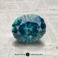 A faceted oval teal 2.11-carat heated Montana sapphire with visible inclusions rests on a textured light gray surface, with "EARTH'S TREASURY" printed at the bottom of the image.