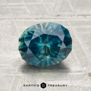A faceted oval teal 2.11-carat heated Montana sapphire with visible inclusions rests on a textured light gray surface, with "EARTH'S TREASURY" printed at the bottom of the image.