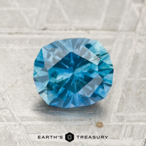 A 1.88-carat heated Montana sapphire, blue and hexagonal, is showcased on a textured gray background with the text "EARTH'S TREASURY" at the bottom.