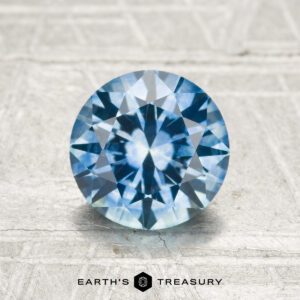 A 1.52-carat heated Montana sapphire, cut in a round brilliant style, rests on a light textured surface with the "EARTH'S TREASURY" logo and text at the bottom.