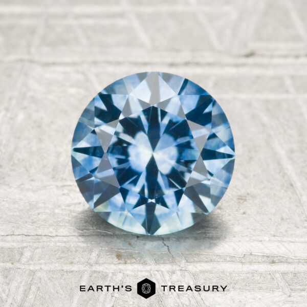 1.52-Carat Montana Sapphire (Heated) A 1.52-carat heated Montana sapphire, cut in a round brilliant style, rests on a light textured surface with the "EARTH'S TREASURY" logo and text at the bottom.