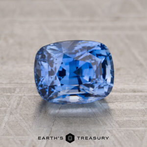 A faceted 2.09-carat Cornflower Blue Sapphire cushion-cut gemstone from "I see the strange light but it seems to come through at every an" rests on a textured gray surface, with "EARTH'S TREASURY" printed beneath.