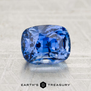 A faceted 2.09-carat Cornflower Blue Sapphire cushion-cut gemstone from "I see the strange light but it seems to come through at every an" rests on a textured gray surface, with "EARTH'S TREASURY" printed beneath.