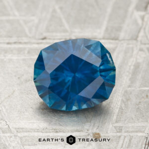 A 1.86-carat heated Montana sapphire with a deep blue hue is showcased on a textured gray surface, with "Earth's Treasury" displayed beneath it.