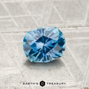 A 0.86-carat heated Montana sapphire is displayed on a textured gray surface with "EARTH'S TREASURY" at the bottom.