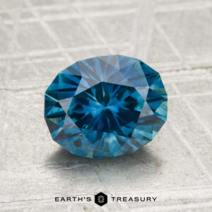 A 2.08-carat heated Montana sapphire, cut in a faceted oval shape, is shown on a textured gray surface with "EARTH'S TREASURY" displayed below.