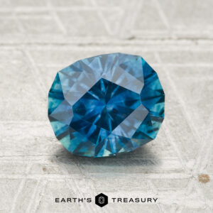 A 1.75-carat heated Montana sapphire gemstone is displayed on a textured pale surface with "EARTH'S TREASURY" printed below.