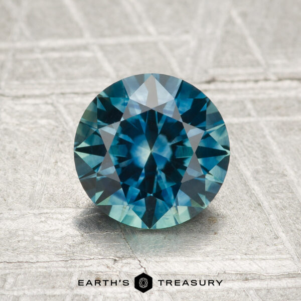 A 1.52-carat heated Montana sapphire, round brilliant-cut and teal in color, is displayed upright on a textured gray surface with "Earth's Treasury" at the bottom.