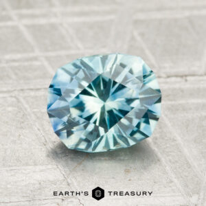 A faceted, light blue 1.69-carat Montana sapphire (heated) is displayed on a textured gray surface with the text "EARTH'S TREASURY" at the bottom.