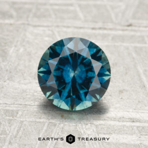 A round, faceted blue-green 1.49-carat Montana Sapphire (heated) is displayed on a textured, light-colored surface, with "EARTH'S TREASURY" at the bottom.