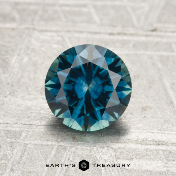 1.49-Carat Montana Sapphire (Heated) A round, faceted blue-green 1.49-carat Montana Sapphire (heated) is displayed on a textured, light-colored surface, with "EARTH'S TREASURY" at the bottom.