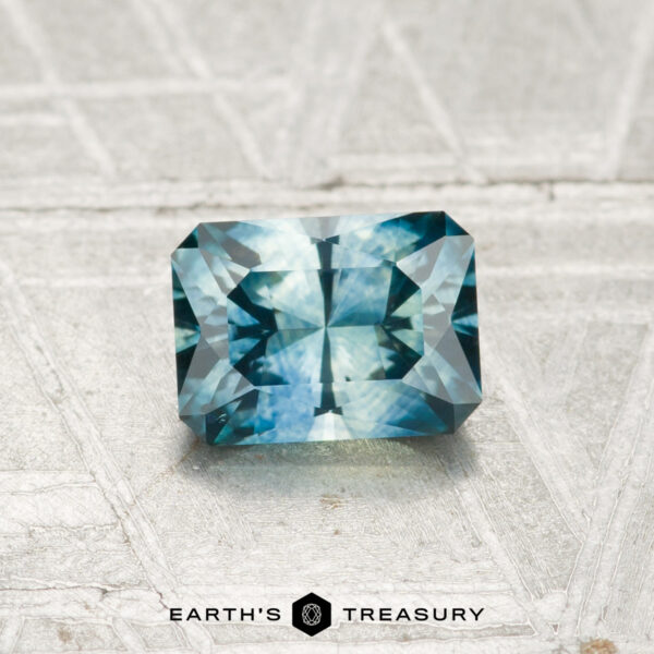 0.86-Carat Montana Sapphire (Heated) A rectangular, faceted 0.86-carat heated Montana sapphire in blue-green rests on a textured light gray surface, with "EARTH'S TREASURY" and two logos visible at the bottom.