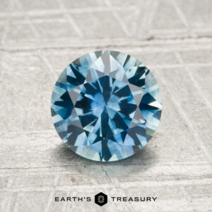 1.48-Carat Montana Sapphire (Heated), round brilliant-cut blue gemstone, showcased on a textured light background with “EARTH’S TREASURY” below.