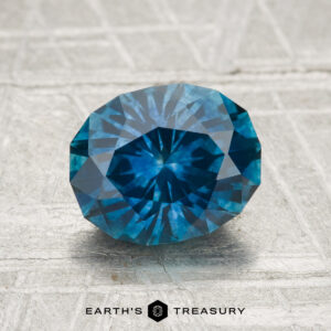 A round, faceted 2.04-carat Montana sapphire (heated) with a blue hue rests on a textured light gray surface. The text "Earth's Treasury" appears below the gemstone.