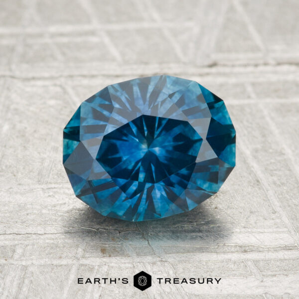2.04-Carat Montana Sapphire (Heated) A round, faceted 2.04-carat Montana sapphire (heated) with a blue hue rests on a textured light gray surface. The text "Earth's Treasury" appears below the gemstone.