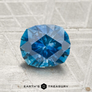 A 1.56-carat heated Montana sapphire with a rich blue color rests on a textured gray surface, “EARTH’S TREASURY” displayed below.