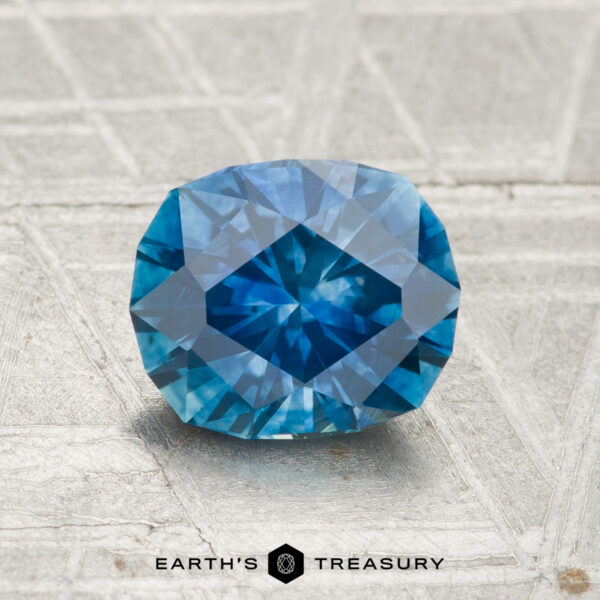 1.56-Carat Montana Sapphire (Heated) A 1.56-carat heated Montana sapphire with a rich blue color rests on a textured gray surface, “EARTH’S TREASURY” displayed below.