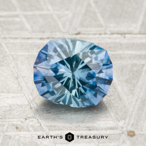 A 1.51-carat heated Montana sapphire, faceted in an oval shape, rests on a textured light gray surface with the “EARTH'S TREASURY” logo below.