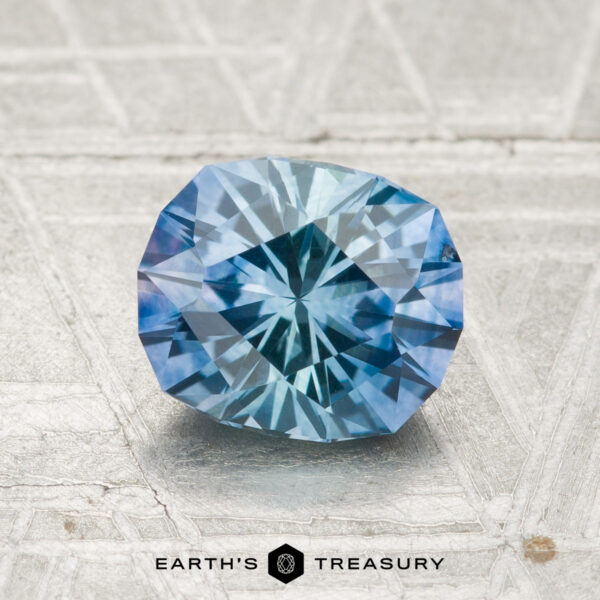 1.51-Carat Montana Sapphire (Heated) A 1.51-carat heated Montana sapphire, faceted in an oval shape, rests on a textured light gray surface with the “EARTH'S TREASURY” logo below.