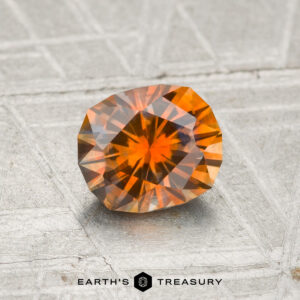 A 1.09-carat heated Montana sapphire with geometric facets rests on a textured, silver-gray surface. "EARTH'S TREASURY" is printed at the bottom of the image.