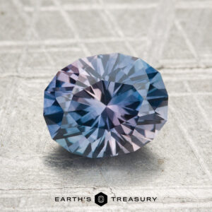 A 2.18-carat heated Montana sapphire with blue and purple hues, cut in a faceted oval shape, rests on a textured surface. "Earth's Treasury" is visible at the bottom of the image.