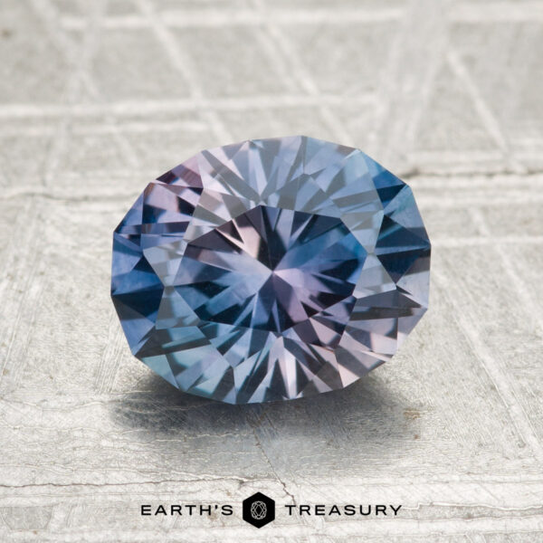 2.18-Carat Montana Sapphire (Heated) A 2.18-carat heated Montana sapphire with blue and purple hues, cut in a faceted oval shape, rests on a textured surface. "Earth's Treasury" is visible at the bottom of the image.