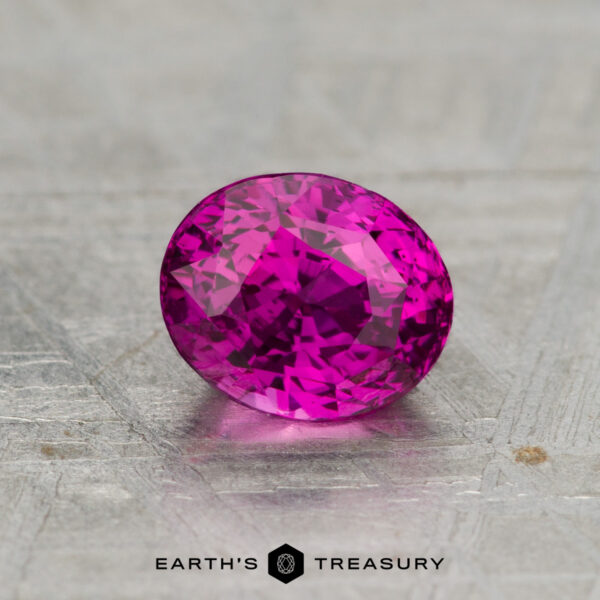 1.33-Carat Sapphire (Heated) A 1.33-carat heated sapphire, faceted in an oval shape and displaying a pink hue, rests on a textured gray surface with "EARTH'S TREASURY" below it.