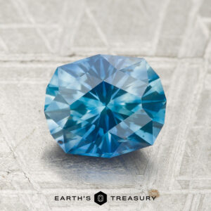 A 2.14-carat heated Montana sapphire with a hexagonal cut is displayed on a metallic surface, with "EARTH'S TREASURY" written below.