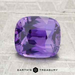 A 3.06-carat Ceylon sapphire with a cushion cut is shown on a textured gray surface, featuring the text "Earth's Treasury" at the bottom.