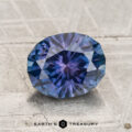 A 2.01-carat heated Montana sapphire with a blue-purple oval cut rests on a textured light gray surface, with "Earth's Treasury" displayed at the bottom.