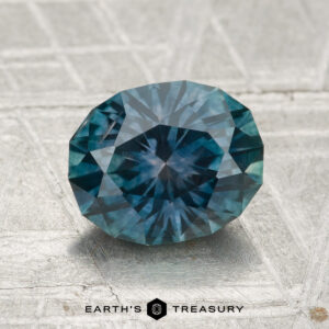A 2.01-carat oval-cut heated Montana sapphire is displayed on a textured gray surface with "EARTH'S TREASURY" beneath it.