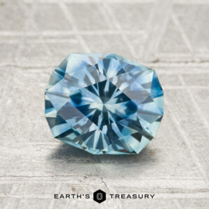 A 1.80-carat heated Montana sapphire with a round brilliant cut is displayed on a textured gray surface, featuring the text "EARTH'S TREASURY" at the bottom.