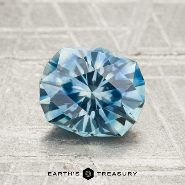1.80-Carat Montana Sapphire (Heated) A 1.80-carat heated Montana sapphire with a round brilliant cut is displayed on a textured gray surface, featuring the text "EARTH'S TREASURY" at the bottom.