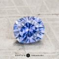 A faceted, oval blue 1.39-carat Montana sapphire (heated) displayed on a textured gray surface with the text "Earth's Treasury" below it.