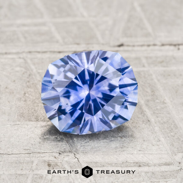 1.39-Carat Montana Sapphire (Heated) A faceted, oval blue 1.39-carat Montana sapphire (heated) displayed on a textured gray surface with the text "Earth's Treasury" below it.