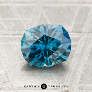 A 1.82-carat heated Montana sapphire, faceted in a blue-green hue, sits on a textured gray surface with "Earth's Treasury" text visible below the gem.