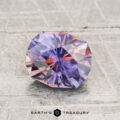 A faceted, oval 1.69-carat heated Montana sapphire with purple and pink hues rests on a textured gray surface, with the text "EARTH'S TREASURY" below.