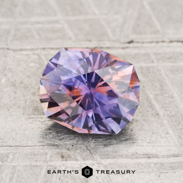 1.69-Carat Montana Sapphire (Heated) A faceted, oval 1.69-carat heated Montana sapphire with purple and pink hues rests on a textured gray surface, with the text "EARTH'S TREASURY" below.