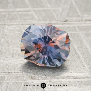 A 1.69-carat heated Montana sapphire with blue and orange hues rests on a textured surface, featuring the "Earth's Treasury" logo at the bottom.
