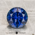 A 2.75-carat heated Montana sapphire in a round brilliant cut rests on a textured light surface, with "Earth's Treasury" displayed beneath.