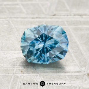 A 1.48-carat heated Montana sapphire is displayed on a textured, light surface with “Earth’s Treasury” below it.