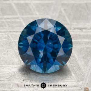 2.75-carat round brilliant-cut heated Montana sapphire displayed on a textured surface, featuring the "Earth's Treasury" logo and text at the bottom.